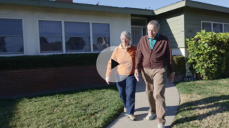 A screenshot of a woman and man exiting their house. They are senior citizens and both white with grey hair. The woman wears a orange sweater, a white button down underneath, and blue jeans. The man wears a brown sweater, a blue button-down underneath, and khakis. They both wear sneakers. They are at the Victory Summit.