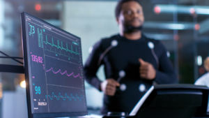 Male Athlete Runs on a Treadmill with Electrodes Attached to His Body while Sport Scientist Holds Tablet and Supervises EKG Status in the Background