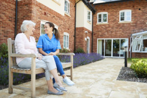 Senior Woman Sitting On Bench And Talking With Nurse In Assisted Living Home