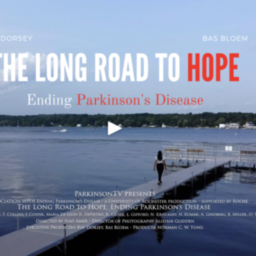 A picture of a dock at a lake with a woman walking down it. It is a screenshot of a film screen title card. The film is called "The Long Road to Hope: Ending Parkinson's Disease", starring Ray Dorsey and Bas Bloem.