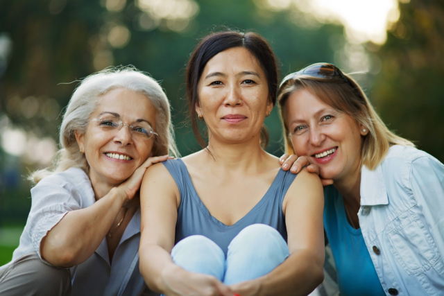 Three women smiling. They are all different ages. One is a white woman with white hair, glasses, and a grey shirt. The middle is an asian woman with black hair, a blue tank top, and light blue pants. The right woman is a white woman with ombre hair in a light blue button-down on top of a cerulean shirt.