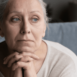 A white woman is sitting, looking like she has demoralization, depression, and meaning with Parkinson's. She has blue eyes, grey hair, and wrinkles. Her hands are clasped under her chin. She is wearing a white shirt on a grey couch.