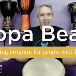 Doug Pickard sits in front of a purple wall covered in bongo drums. He is a white man with short, grey hair. Super-imposed on him is the word "DopaBeats" in white. Below that is a yellow bar wtih the words "a drumming program for people with Parkinson's". The Davis Phinney Foundation logo is in the bottom left corner.