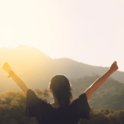 Parkinson's optimism. A white woman stands with her back to the camera. She is holding up victory arms before a forested mountain landscape. She has a brown ponytail and a black shirt.