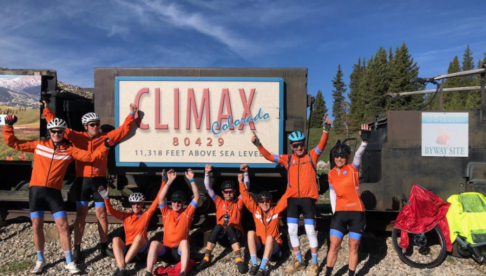 Sarah Zenner and her team share their Moments of Victory at the Climax of Colorado. They are using Davis Phinney's Victory Arms. They are wearing orange bike jerseys, bike pants, and helmets. There is bike on the left side of the picture.