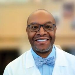 Parkinson's disease psychosis. Dr. Dylan Wint, a black man, sits in front of a blurry office background. He is wearing a white lab coat, a blue button down, a blue bowtie, and black glasses. He smiles with teeth.