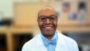 Parkinson's disease psychosis. Dr. Dylan Wint, a black man, sits in front of a blurry office background. He is wearing a white lab coat, a blue button down, a blue bowtie, and black glasses. He smiles with teeth.