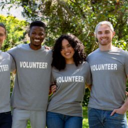 Four volunteers who live well with Parkinson's. They are a white man with black hair, a black man with black hair, a brown woman with curly, shoulder-length brown hair, and a white man with a brown beard and silver hair. They are all wearing jeans and grey shirts that say "volunteer".