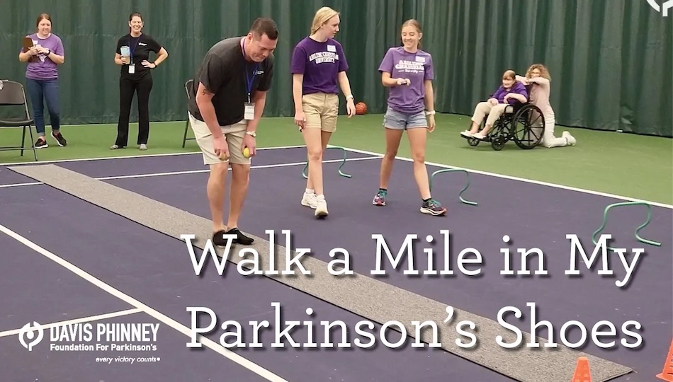 Walk a Mile in My Parkinson's Shoes - Davis Phinney Foundation lorraine wilson parkinson's Empathy—a candid photo of an indoor track with various walkers on it. The track is purple, and a green floor is next to it. The curtains are green. Seven people in the photo wear a mix of purple and black shirts. There is a person in a wheelchair in the background.