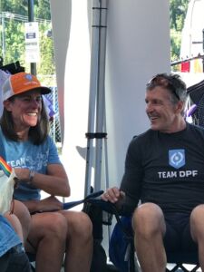 Sarah Zenner, in a blue Team DPF shirt and a orange and white Team DPF trucker hat sits in a lawn chair next to Davis Phinney, who is wearing a black Team DPF shirt and black shorts. Sarah has shoulder-length brown hair and Davis has short grey hair with biking sunglasses on his head. They are both smiling.