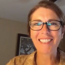 Parkinson's depression. Joanne hamilton is smiling and wearing glasses. She has brown hair and a yellow button-down. She is in front of a room with a fan.