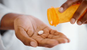 a person is giving themselves rasagiline. It is a black woman's hands with a single pill in her right hand and a pill bottle in the other.