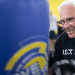 William Richard stands at a punching bag. He is a white senior citizen man with a black shirt and white hair and glasses.