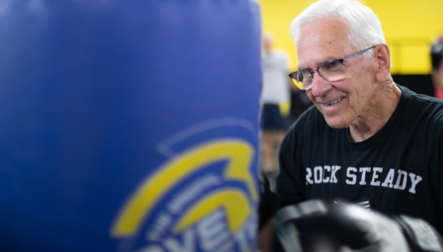 William Richard stands at a punching bag. He is a white senior citizen man with a black shirt and white hair and glasses.