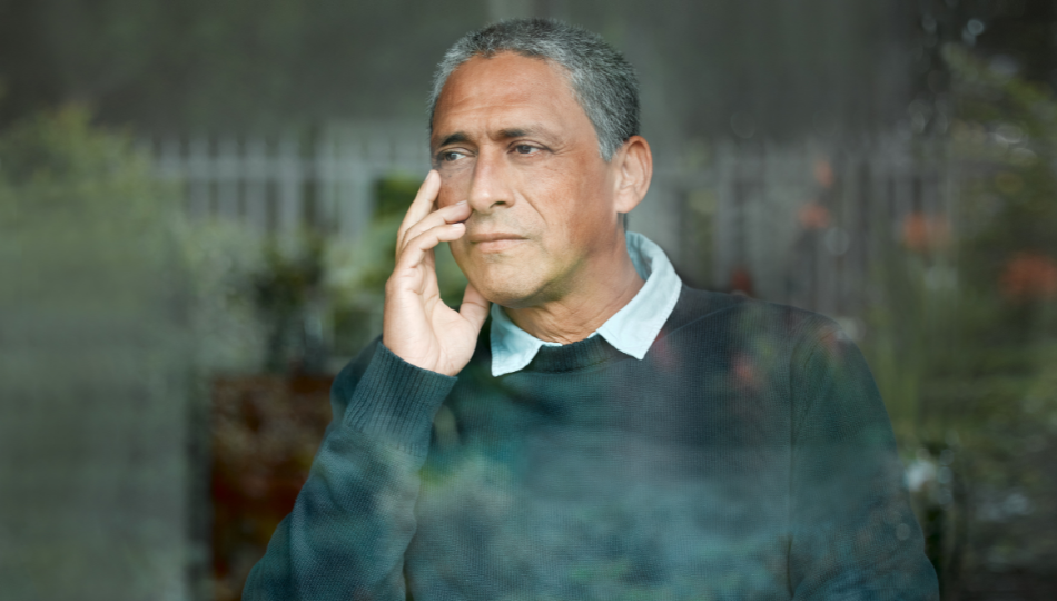 A confused Black man is holding a phone to his right ear. He is staring out a clear window, though we can see a small reflection of what looks like plants outside. He is wearing a grey sweater with a white button-down underneath. His hair is greying, curly, and short.