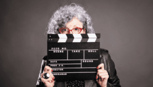 Grey, Curly haired woman is holding a clapper in front of her face, only showing her red glasses. She is wearing a leather jacket, has red nail polish, and a large, silver ring on her right hand.