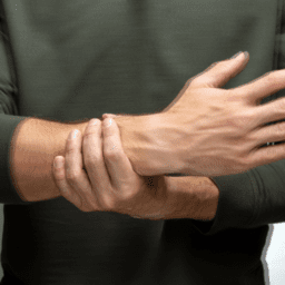 Man holding wrist. Only his torso is showing. He is wearing a dark green/grey shirt. He has white skin.