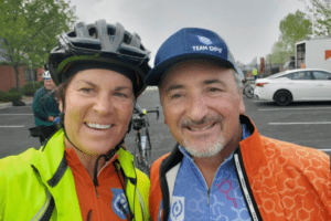 Ellen Nordberg, wearing a black helmet, red bike jacket, and bright yellow over jacket, smiles with her teeth next to Tom Palizzi who is wearing a Team DPF hat, a orange biking jacket, and a blue DPF bike jersey.