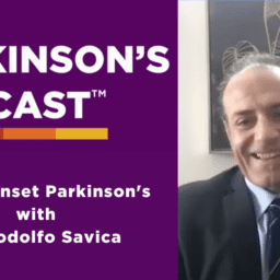 The Parkinson's Podcast logo with the words "Young Onset Parkinson's with Dr. Rodolfo Savica" is on the left side of the picture. The right side of the picture is a picture of Rodolfo Savica. He is in front of a black abstract painting and white wall. He is a balding, white man, wearing a charcoal suit, a blue shirt, and a striped navy tie.