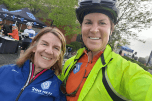 Polly Dawkins and Ellen Nordland stand next to each other. Ellen is taking the selfie. Polly is wearing a blue quarter zip. Her hair, honey brown, is shoulder-length and straight. Ellen is wearing a black helmet, a neon yellow overcoat, and an orange bike jersey.
