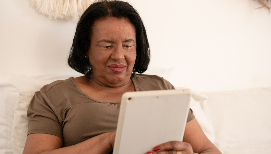 A black woman, middle-aged, looks at an iPad. She has chin-length black hair, red lipstick, and a black shirt. Her iPad case is white. She has red nail polish and is sitting on a white couch with a white wall behind her.