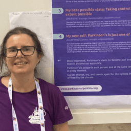 Lucia Wang, a brown-haired woman with a purple shirt and black glasses, stands in front of a blue and white poster. The poster has words about Parkinson's at the 6th World Parkinson's Congress in Barcelona, Spain.