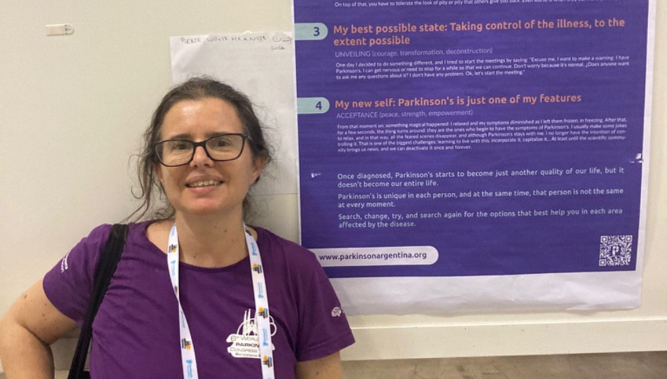 Lucia Wang, a brown-haired woman with a purple shirt and black glasses, stands in front of a blue and white poster. The poster has words about Parkinson's at the 6th World Parkinson's Congress in Barcelona, Spain.