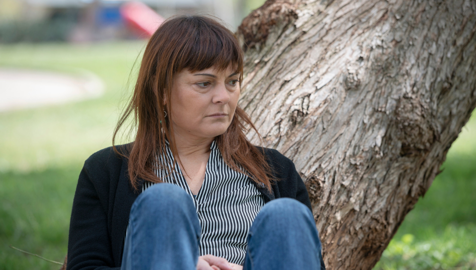 A white woman with shoulder-length brown hair and bangs is sitting in front of a tilted tree. She looks contemplative and sad. She is wearing a black cardigan, a vertical-striped black and white button-down, and blue jeans. There is a playground in the background.