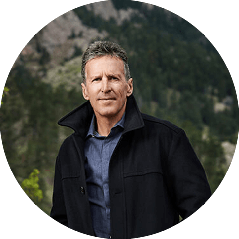 Davis Phinney stands in front of a mountain covered in trees. He is wearing a black coat and a denim button-down. He has short, grey hair and a stoic look on his face.