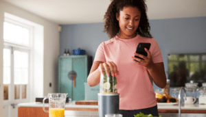 young black woman looking at her phone. She is learning to track her symptoms. She is blending a cucumber smoothly. She is in her kitchen. She is wearing a pink shirt.
