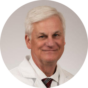 Mark Stacy, MD, a white man with white hair. he is giving a half smile. he is wearing a white lab coat with the MUSC Health logo, a white button-down, and a red tie with blue stripes. 