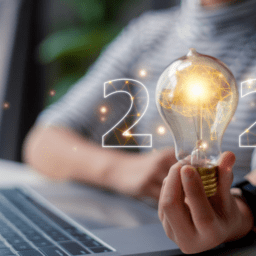 free a person holding a light bulb in front of a laptop with the number 2024 written in front of the blurry background. The 0 is the light bulb. There are lines and dots that are glowing gold throughout the picture. The lightbulb has a brain in it.