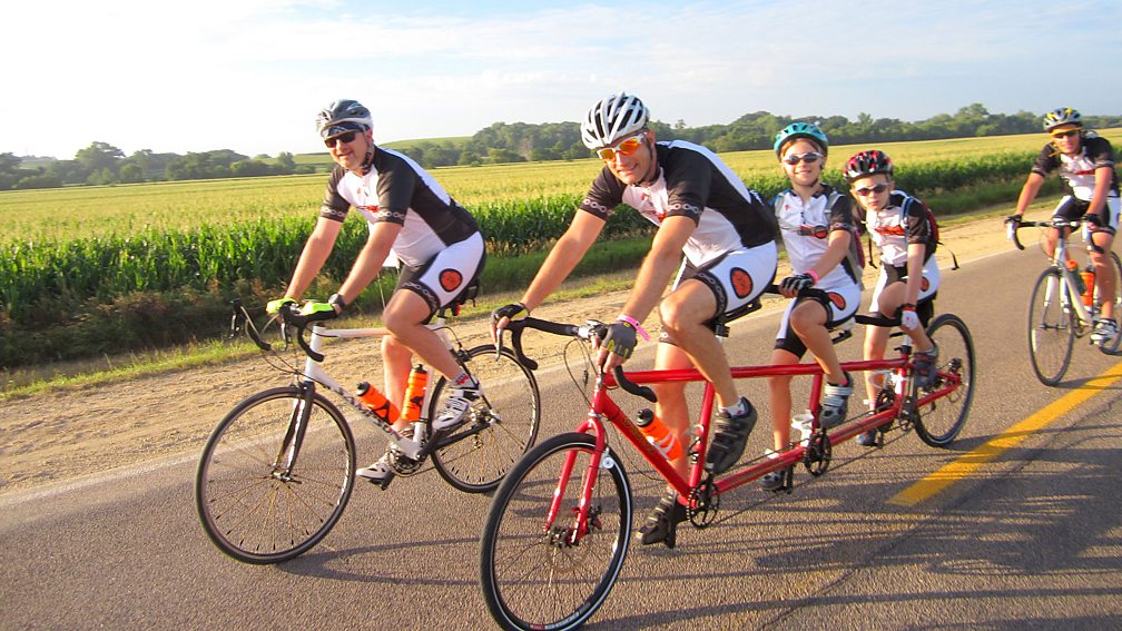 Five cyclists wearing Pedaling for Parkinson's gear riding on a road next to an open field. In the foreground is neuroscientist and PFP co-founder, Dr. Jay Alberts, on a 3 seater bike with his two kids in Iowa.