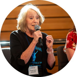 Carolyn Utesch in a "BadAssadors" shirt, speaking into a microphone. Carolyn is a middle-aged white woman with blonde hair.