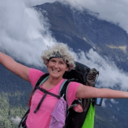 Kristin Matteson has her arms spread wide as she climbs a mountain, with a mountain range visible behind her. She is smiling with her white hair, pink shirt, and green and black backpack.
