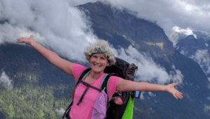 Kristin Matteson has her arms spread wide as she climbs a mountain, with a mountain range visible behind her. She is smiling with her white hair, pink shirt, and green and black backpack.